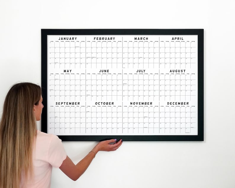 May include: A large, framed wall calendar with a black frame. The calendar grid is white with black text, displaying the months of the year. The person is holding the calendar against a white wall.