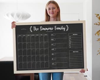 May include: A large rectangular wall calendar with a black chalkboard surface and a light wood frame. The board features a monthly grid, a weekly schedule section, a menu planner, and a notes section, all printed in white text with The Simmons Family.