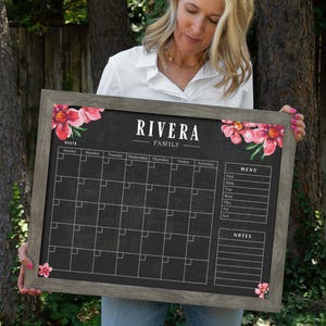 以下が含まれることがあります： 花柄のデザインと「RIVERA FAMILY」の文字が書かれた灰色の額縁の黒板。黒板には月間カレンダー、メニュー欄、メモ欄があります。