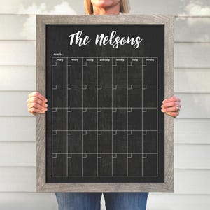 May include: A black chalkboard with a wood frame, labeled "The Nelsons" at the top. The chalkboard has a monthly calendar grid with days of the week labeled.