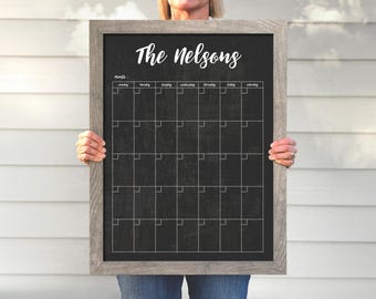 May include: A black chalkboard with a wood frame, labeled "The Nelsons" at the top. The chalkboard has a monthly calendar grid with days of the week labeled.
