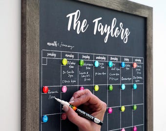 May include: A framed chalkboard calendar with the text "The Taylors" in white script. The calendar grid is filled with colorful magnets and handwritten appointments for January. A hand is writing on the board with a white marker.
