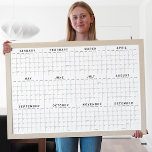 May include: A large, framed wall calendar with a light wood frame. The calendar is white with black text, displaying the months of the year. The calendar is held by a person.