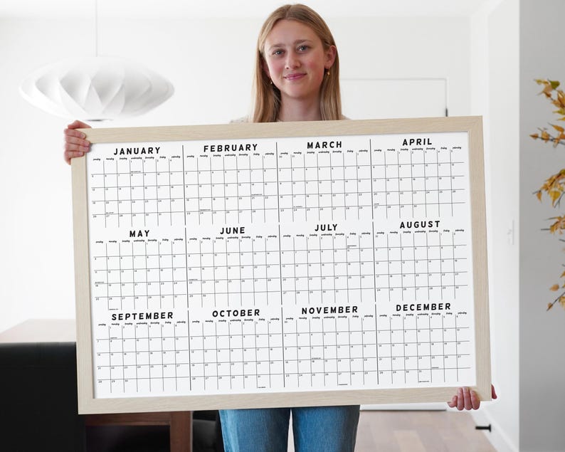 May include: A large, framed wall calendar with a light wood frame. The calendar is white with black text, displaying the months of the year. The calendar is held by a person.