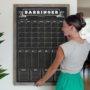 以下が含まれることがあります： 「Barrringer」と書いた、額縁に入った黒板の月間カレンダー。カレンダーは、月の各日に対応する正方形のグリッドになっており、上部には曜日がラベル付けされています。下部には、「Ruby」、「Ben」、「Riley」などの名前のセクションもあります。