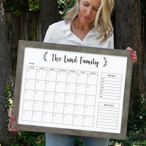 May include: A white dry-erase calendar with a gray wood frame. The calendar has a monthly grid with spaces for each day of the week. It also has sections for notes and a menu. The text "The Lund Family" is written at the top of the calendar.