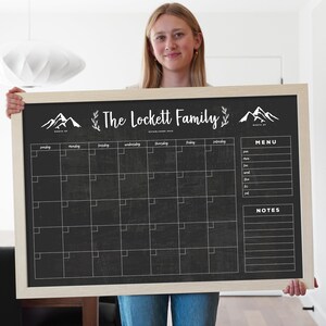 May include: A large, rectangular chalkboard calendar with a light wood frame. The black surface features a grid for dates, a menu section, and a notes section. The top of the board displays the text "The Lockett Family" with mountain and floral graphics.