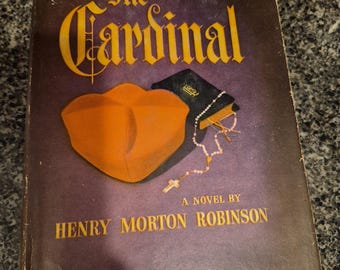 Könnte beinhalten: Vintage Buchcover für "The Cardinal" von Henry Morton Robinson. Das Cover zeigt einen goldenen Titel und Autorennamen auf dunklem lila Hintergrund. Ein orangefarbener Hut und ein kleines Buch mit Rosenkranz sind ebenfalls abgebildet.