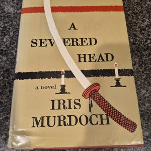 Może przedstawiać: Vintage okładka książki "A Severed Head" autorstwa Iris Murdoch. Okładka przedstawia stylizowaną katanę z czerwonym uchwytem i białym ostrzem, na beżowym tle z czarnym tekstem i czerwonym paskiem. Obecne są również dwie ilustracje świec.