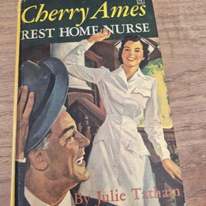 May include: Vintage paperback book titled "Cherry Ames Rest Home Nurse" by Julie Tatham. The cover features an illustration of a smiling nurse in a white uniform and a man in a suit and hat. The book has a green and yellow cover.