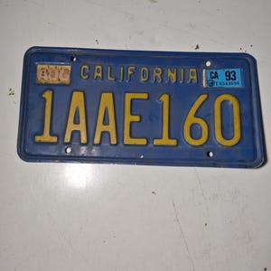 Pode incluir: Uma placa de carro da Califórnia vintage com fundo azul e letras amarelas. A placa diz "1AAE160" e "CALIFORNIA". Um adesivo no canto superior direito indica "CA 93". A placa mostra sinais de desgaste.