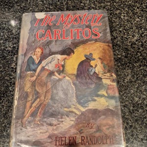 May include: Vintage hardcover book titled "The Mystery of Carlitos" by Helen Randolph. The cover features an illustration of two people in a cave, with a figure in a hat and a table. The title is in red, with a faded, aged appearance.