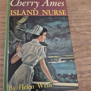 Può includere: Copertina di libro vintage di "Cherry Ames Island Nurse" di Helen Wells. La copertina presenta un'infermiera che guarda l'oceano. Il titolo è in lettere grandi e in grassetto con un dorso verde.