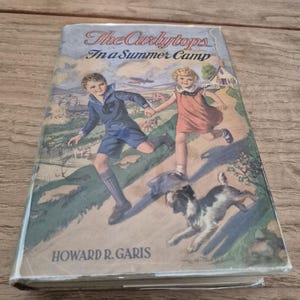 May include: Vintage hardcover book titled "The Curlytops In a Summer Camp" by Howard R. Garis. The cover features a colorful illustration of two children running with a dog, set against a summer camp backdrop.