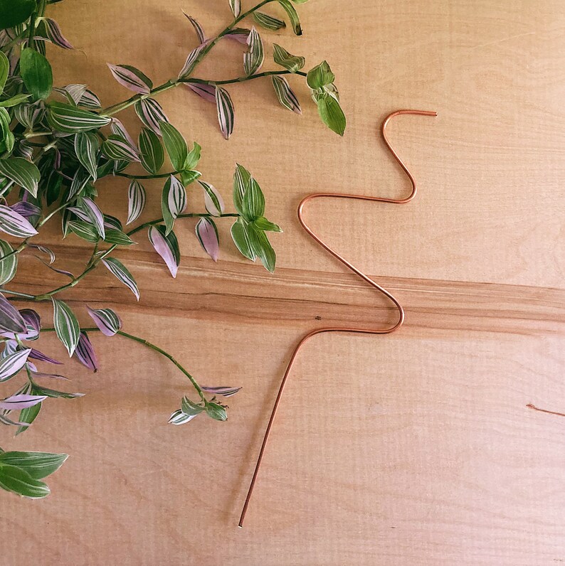 May include: A copper wire plant stake with a wavy design. The stake is on a light brown wooden surface.