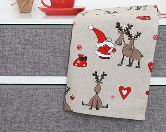May include: A beige kitchen towel with a Christmas theme. The towel features a red and white Santa Claus, reindeer, and snowflakes. The towel is folded and placed on a gray countertop.