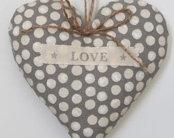 May include: A gray heart-shaped fabric ornament with white polka dots. A beige ribbon with the word "LOVE" is attached. The ornament is suspended by a twine string and a decorative crystal.