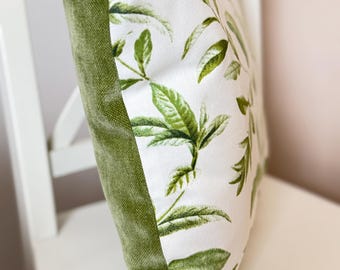 May include: A decorative pillow with a white background and a green leaf pattern. The pillow has a green velvet border. The pillow is resting on a white chair.