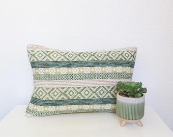 May include: A rectangular decorative pillow with a geometric pattern in shades of green, cream, and blue. The pillow is placed on a white surface next to a small green and white potted plant. The pillow measures approximately 20 inches long.