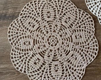 May include: Beige crocheted doilies with a floral design. The doilies are circular with a detailed openwork pattern. The crochet work is visible, and the doilies are placed on a wooden surface.