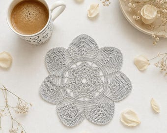 May include: A light gray, circular crochet doily featuring a detailed, intricate floral pattern with six rounded petals radiating from a central open ring. The piece is crafted with fine thread, showcasing a delicate, openwork stitch design.