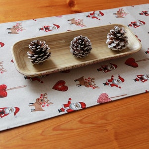 May include: A beige table runner with a repeating pattern of red hearts, reindeer, and Santa Claus figures. A wooden tray with three pine cones sits on top of the runner.