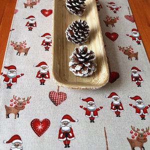 May include: A beige table runner with a Christmas theme featuring red and white hearts, reindeer, and Santa Claus figures. Three pine cones are displayed in a wooden tray.