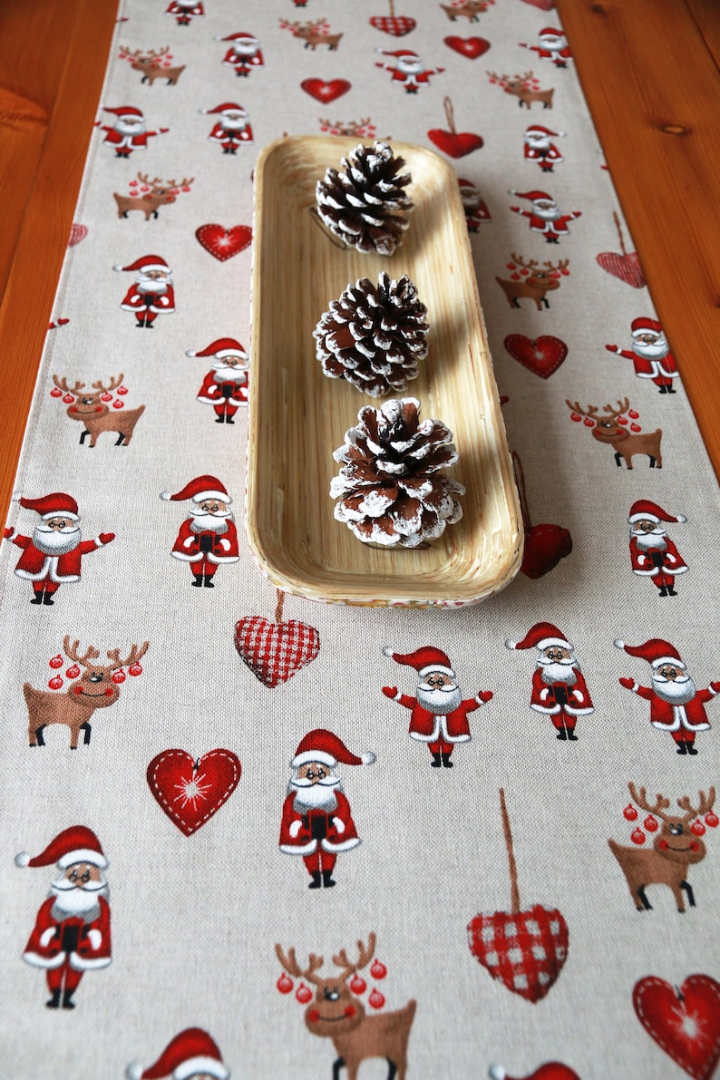 May include: A beige table runner with a Christmas theme featuring red and white hearts, reindeer, and Santa Claus figures. Three pine cones are displayed in a wooden tray.