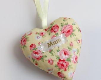 May include: A heart-shaped fabric ornament with a floral pattern of pink roses on a yellow background. The word "Mum" is printed on a white ribbon. A light green ribbon is attached for hanging.