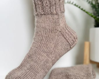 May include: A pair of knitted socks in a light brown color. One sock is displayed on a mannequin foot, while the other lies beside it. The socks have a ribbed cuff and a textured knit pattern throughout the body.