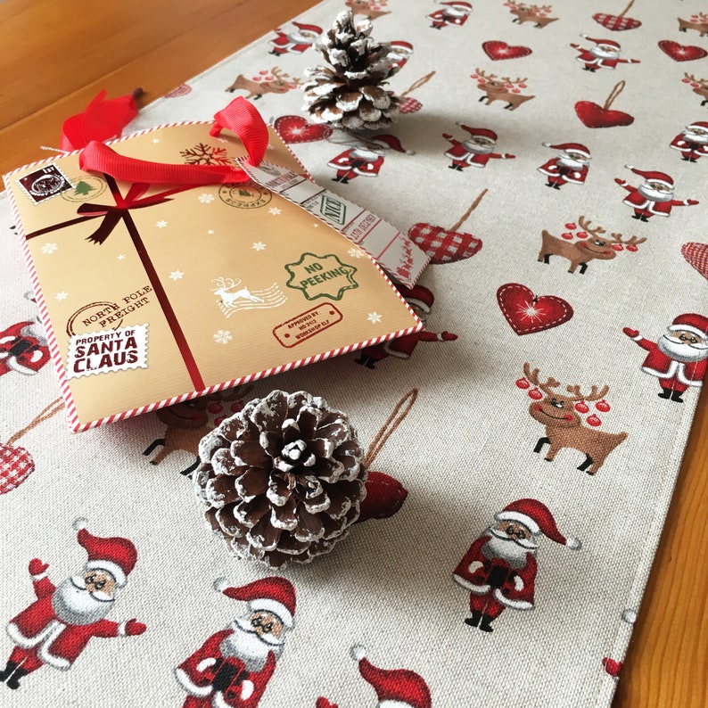 May include: A beige table runner with a festive Christmas theme. It features a repeating pattern of red hearts, reindeer, Santa Claus figures, and pine cones. The text "North Pole Freight" and "Property of Santa Claus" is printed on a brown paper package with red ribbon.