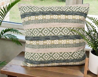 May include: A square decorative pillow with a geometric pattern in shades of green, beige, and dark teal. The pillow is placed on a wooden surface near a window with plants. The design features diamond and stripe patterns.