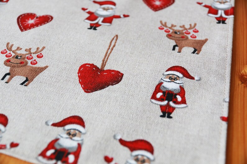 May include: A beige fabric table runner with a repeating pattern of red hearts, Santa Claus figures, and reindeer with red ornaments. The pattern is festive and perfect for the Christmas season.