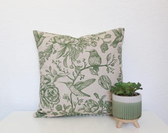 May include: Square throw pillow with a botanical design featuring hummingbirds and flowers in green on a beige background. A small green and white potted plant sits next to the pillow.