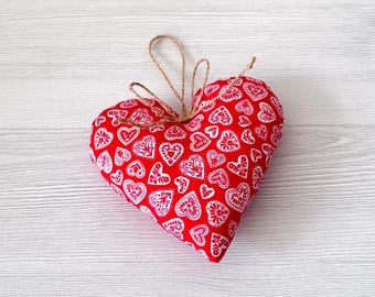 May include: A red heart-shaped fabric ornament with a white floral pattern. The ornament has a twine loop for hanging. The heart is filled with stuffing and is placed on a light wood grain surface.