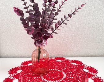 May include: A round, bright red crocheted doily with an intricate, openwork floral pattern. A clear, ribbed glass vase sits in the center, holding several stems of artificial eucalyptus branches with deep purple, rounded leaves.
