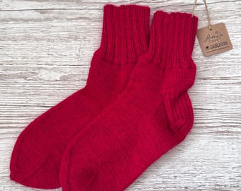 May include: A pair of bright red knitted socks with a ribbed cuff design. A small brown cardboard tag is attached to the top of one sock with a string, featuring the printed text Arda Da CRAFT and various laundry care symbols.