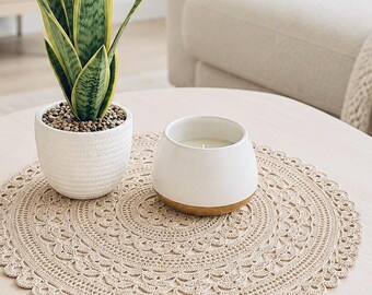 May include: A round beige crochet doily with an intricate lace pattern featuring scalloped edges. A white ceramic planter with a green and yellow snake plant and a white candle in a bowl with a wooden base sit on top of the textured fabric.