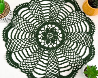 May include: A round, dark forest green crochet doily featuring an intricate, openwork floral pattern that radiates from a central star-like motif toward a scalloped edge. The design consists of repeating mesh segments and delicate yarn loops.