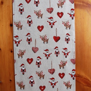 May include: A beige table runner with a repeating pattern of red hearts, reindeer, and Santa Claus figures. The runner is made of a linen-like fabric.