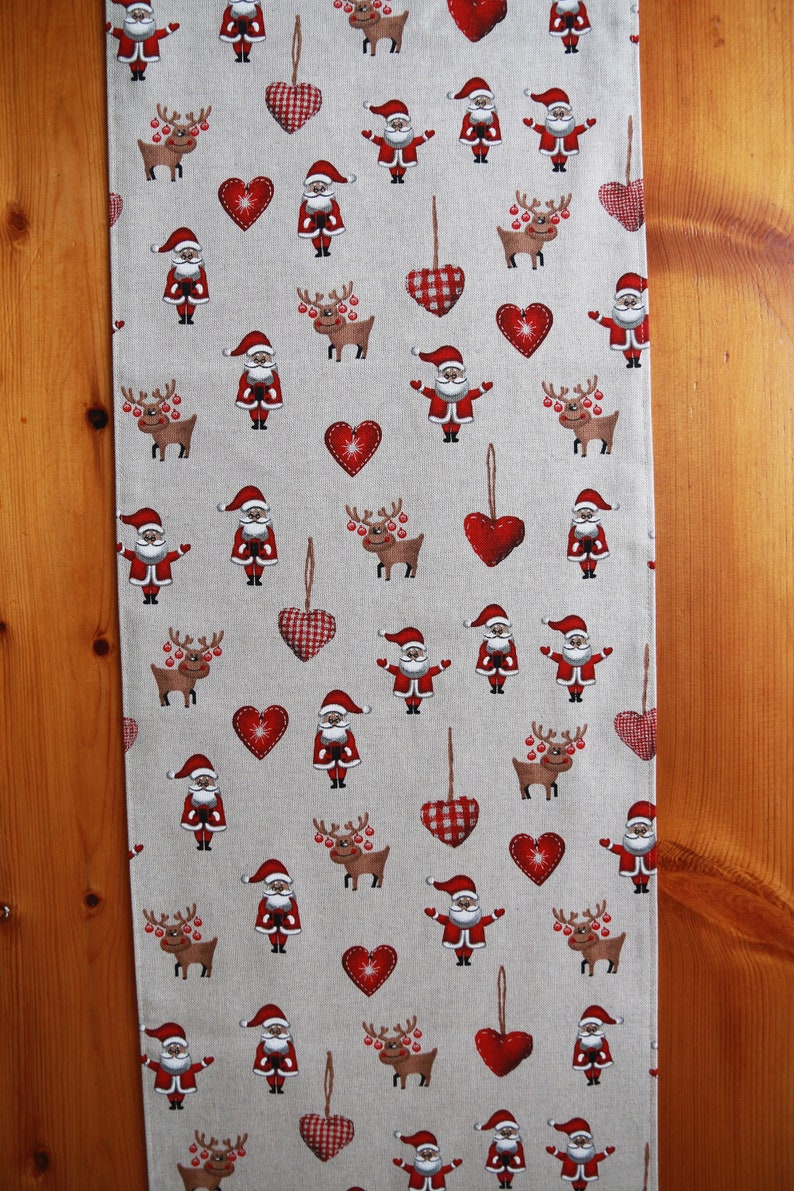 May include: A beige table runner with a repeating pattern of red hearts, reindeer, and Santa Claus figures. The runner is made of a linen-like fabric.