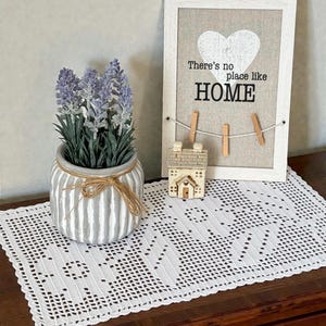 May include: A white crocheted rectangular doily, a grey and white striped ceramic pot with purple lavender, a small beige house figurine, and a white framed sign with a heart and the text There's no place like HOME with three wooden clips.