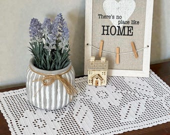 May include: A white crocheted rectangular doily, a grey and white striped ceramic pot with purple lavender, a small beige house figurine, and a white framed sign with a heart and the text There's no place like HOME with three wooden clips.