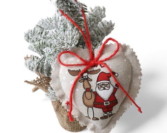 May include: A heart-shaped Christmas ornament with a Santa Claus and reindeer design, tied with a red string. The ornament is made of beige fabric and is attached to a small, snow-covered Christmas tree in a burlap pot.