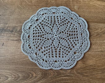 May include: A round, gray crocheted doily with a scalloped edge. The intricate openwork design features a central floral pattern and radiating lines. This handmade item is suitable for home decor.