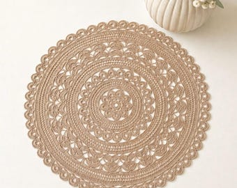 May include: A round, beige crochet doily featuring a central floral motif surrounded by concentric rings of intricate, openwork stitch patterns and a scalloped edge. The design is composed of fine thread, creating a detailed, circular texture.