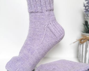 May include: A pair of hand-knitted lavender socks featuring a ribbed cuff and a smooth knit foot section. The socks are crafted from a soft, heathered yarn, with one sock displayed upright and the other folded neatly beside it.