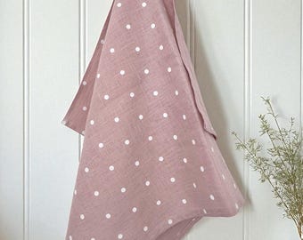 May include: A dusty rose-colored kitchen towel with white polka dots hangs from a wooden bar. The towel is made of linen and has a loop for hanging. The background is a white wall with vertical paneling.