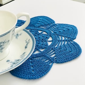 May include: A blue crocheted doily with a delicate floral pattern sits on a white table with a white teacup and saucer.