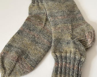 May include: A pair of hand-knitted socks in muted tones of green, gray, and brown. The socks have a ribbed cuff and a textured, speckled appearance, suggesting a natural fiber yarn. The socks are designed for warmth and comfort.